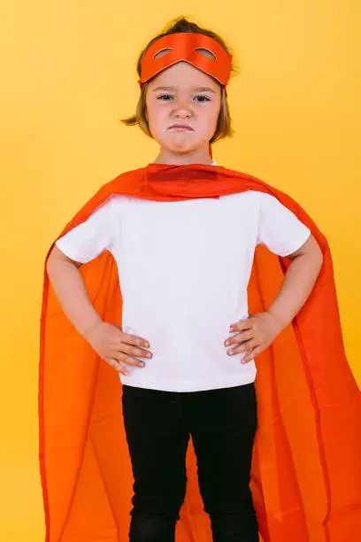 Little blonde girl dressed as a superheroine superhero with a cape and red mask, angry, with her arms akimbo serious, on yellow background