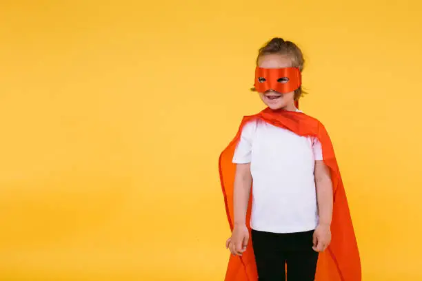 Little blonde girl dressed as superheroine superhero with cape and red mask, smiling and looking to the side, on yellow background