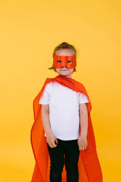 Little blonde girl dressed as superheroine superhero with cape and red mask, smiling and looking at camera, on yellow background