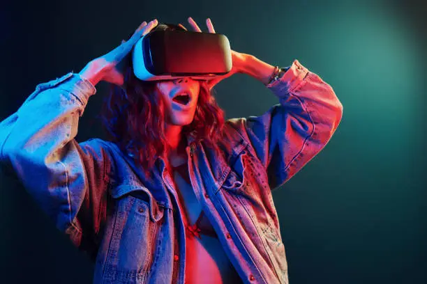 Facial expression of young girl with virtual reality glasses on head in red and blue neon in studio.