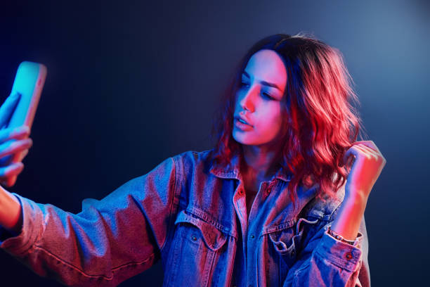 retrato de una joven con el pelo rizado que se hace selfie usando el teléfono en neón rojo y azul en el estudio - falso fotos fotografías e imágenes de stock