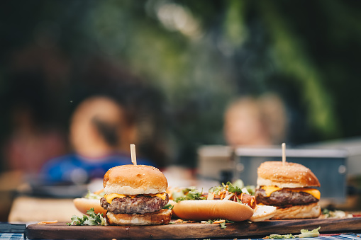 Two hamburgers with beef burger cutlet and blue cheese in traditional buns, served on wood chopping board. Hot dogs are almost reedy and chef has arranged fresh food for guests.