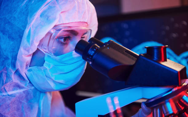 istock Nurse in mask and white uniform sitting in neon lighted laboratory with computer, microscope and medical equipment searching for Coronavirus vaccine 1324023302