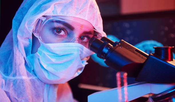 istock Nurse in mask and white uniform sitting in neon lighted laboratory with computer, microscope and medical equipment searching for Coronavirus vaccine 1324023328