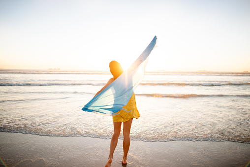 Rear view of a young woman holding a scarf blowing in the wind while watching the sunset on a sandy beach