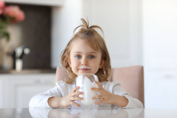 blond cute baby girl with blue eyes with traces of milk on the lips is holding a glass of milk siting at a white table in a white kitchen. milk for good health - milk mustache imagens e fotografias de stock