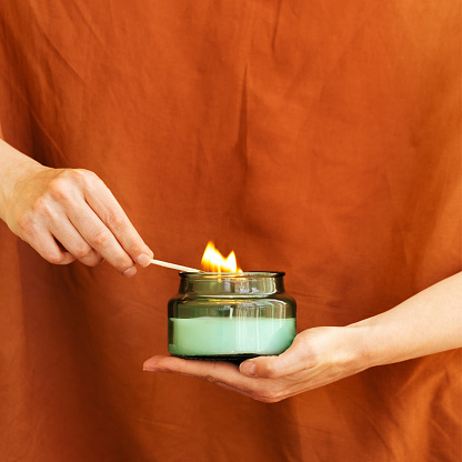 Womans hand uses match to light aromatic candle in recycled glass jar on terracotta colored background. Wellness and physical, emotional health concept.