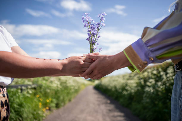 A side-view shot of an unrecognizable LGBTQIA female couple standing on a footpath together, they are wearing casual clothing, they're holding hands and flowers together on a romantic day out.