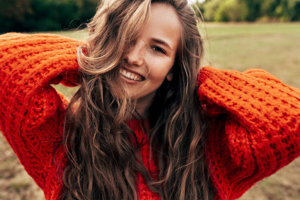 Outdoor portrait of a smiling young woman wearing a knitted orange sweater posing on nature background. The beautiful female has a joyful expression, resting in the park. stock photo