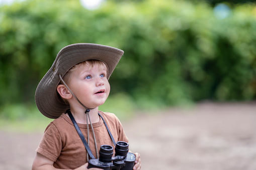 A three year old Caucasian boy holds a pair of binoculars while exploring the outdoors.