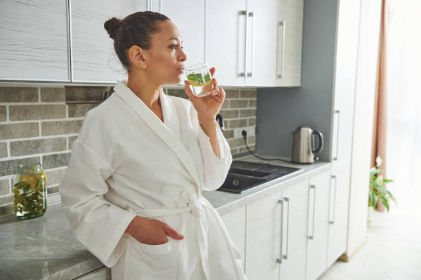 Cute woman in a white robe, leading a healthy lifestyle, drinking lemon water leaning against the kitchen countertop. A beautiful woman in a white robe, leading a healthy lifestyle, drinking lemon water leaning against the kitchen countertop. Morning started with warm lemon water detox stock pictures, royalty-free photos & images