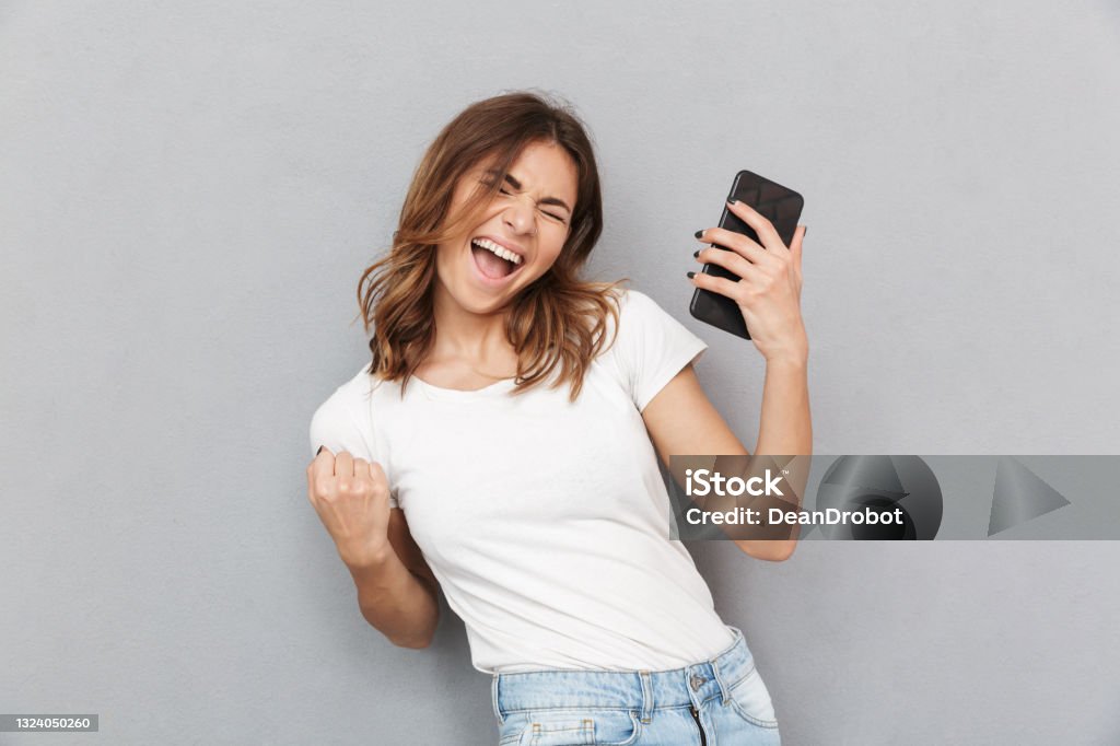 Portrait of a satisfied young woman Portrait of a satisfied young woman holding mobile phone and celebrating over gray background Happiness Stock Photo
