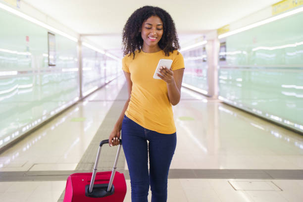 una mujer negra que viaja con teléfono inteligente y equipaje de mano - airport business travel arrival departure board travel fotografías e imágenes de stock