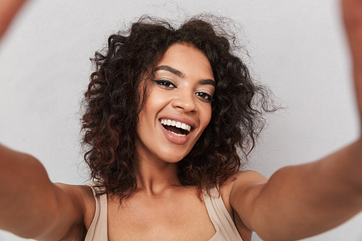 Close up portrait of a smiling young african woman taking a selfie with outstretched arms over gray background