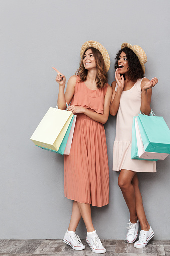 Full length portrait of two happy young women dressed in summer clothes holding shopping bags and looking away over gray background