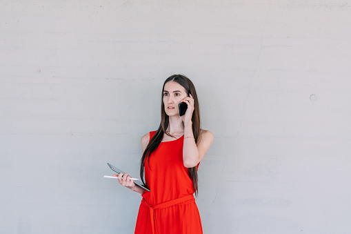 Young business woman in red dress, holding a tablet while receiving a phone call with her cell phone.