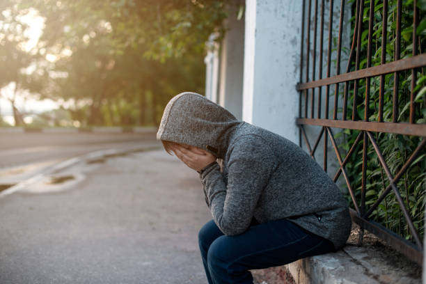 A lonely and sad teenage student with his face covered with his hands, in a closed pose hiding from problems on the street in an urban environment. The concept of social problems of youth. stock photo