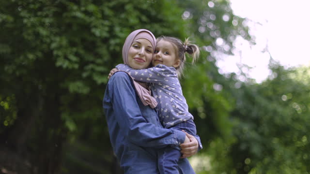 Slow motion of pleasant pretty young Muslim mother wearing casual jeans clothes and beige hijab, holding on hands her cute little daughter and hugging each other while walking at park.