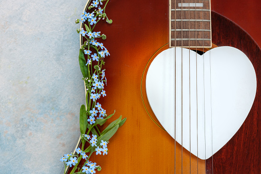 Acoustic guitar, wooden white heart shape symbol and forget-me-not flowers on sky blue background. Top view, close up, copy space.