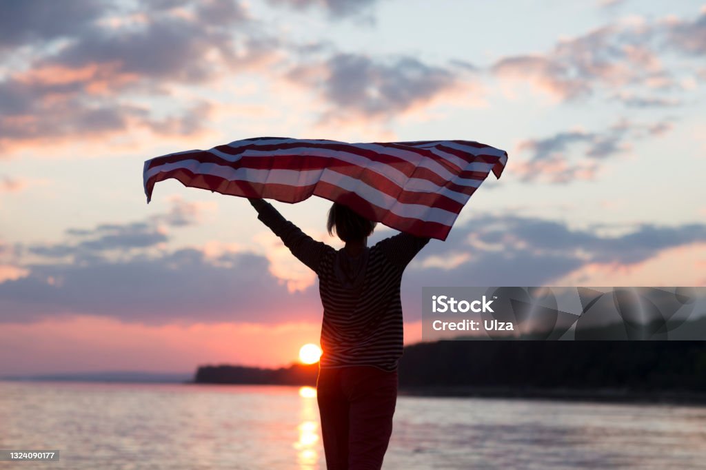a woman with an American flag on the ocean at sunset. Independence Day. The United States celebrates July 4. American Flag Stock Photo