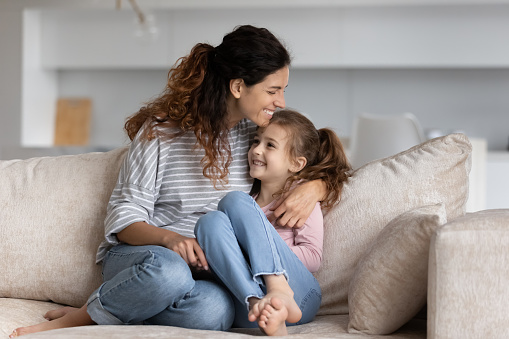 Happy Latino young mother and small teen daughter sit relax on sofa at home have fun hug cuddle. Smiling Hispanic mom and little girl child embrace enjoy family weekend together. Bonding concept.