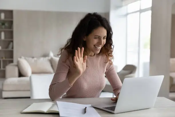 Smiling Hispanic woman work online on computer at home greet wave talk speak on video call on device. Happy Latino female use laptop have webcam video virtual event with client or teacher.