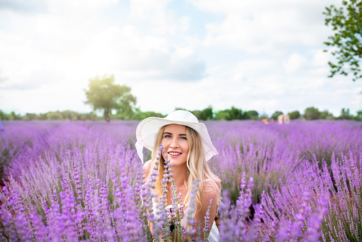 Young beautiful woman in white dress and white hat walking in lavender field.
Beautiful woman smells lavender flowers.