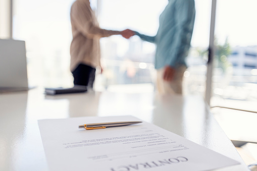 Two businessmen shaking hands with a contract. There is a pen on the contract document. Focus is on the foreground with the people in the office out of focus in the background. They are wearing casual business clothes.