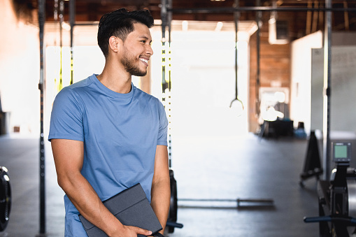 The mid adult male fitness instructor smiles at an unseen person at the gym.  He is holding his digital tablet.