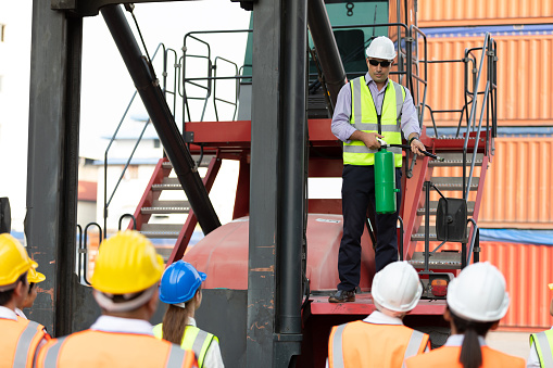 male engineer standing on crane car using fire extinguisher with factory workers in containers warehouse storage