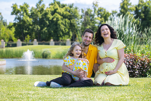 Family portrait in the park. Two parents with their daughter are sitting on the grass near the lake. Father holds his daughter in his arms. The mother is pregnant with her second child at the last term.