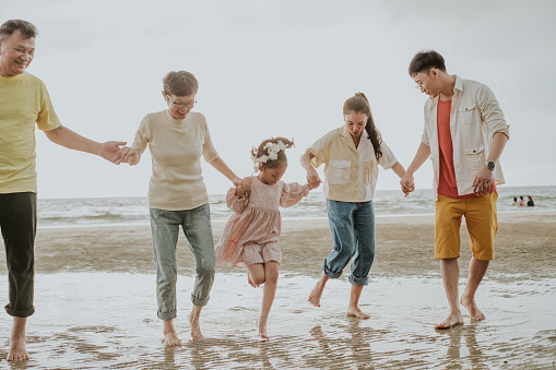 Unity family who love to travel the sea together, multi-generation of Southeast Asian family holding hands together while resting thee beach for the long weekend