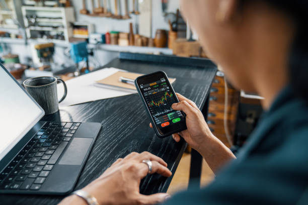 Mid adult man using a smart phone to monitor his cryptocurrency and stock trading Mid adult man using a smart phone to monitor his cryptocurrency and stock trading. He is in his small jewellery workshop. stock exchange stock pictures, royalty-free photos & images