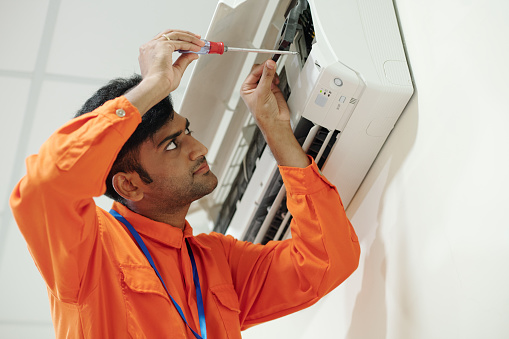Concentrated young Indian engineer in orange wear using screwdriver while setting up air conditioner