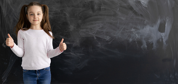 a Caucasian girl, a schoolgirl, is standing against the background of an empty chalk board. The thumb is raised up. copy space.