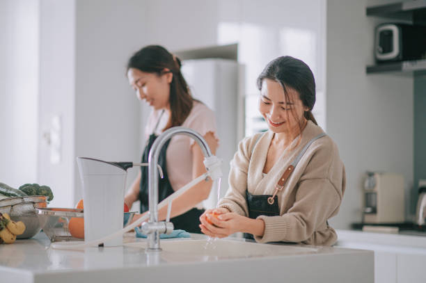 asian chinese female hand washing cleaning orange fruit at sink with running water at home during weekend at kitchen counter using laptop - team human hand cheerful close up imagens e fotografias de stock