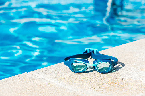 Blue swimming goggles on the curb of the pool, in the lower right corner, with the blue water in the background