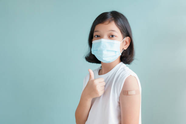Asian little girl wearing face mask vaccinated Gesturing Thumbs Up  Showing arm bandage to protect COVID-19 spread on blue background stock photo