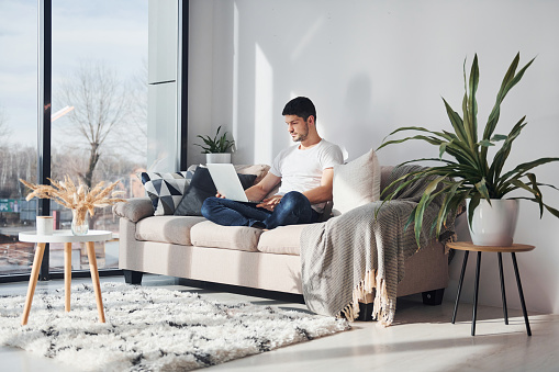 Man in white shirt and jeans sitting on bed with laptop indoors.