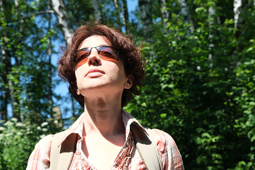 Mature woman wearing sunglasses is standing under the trees on a hot sunny day