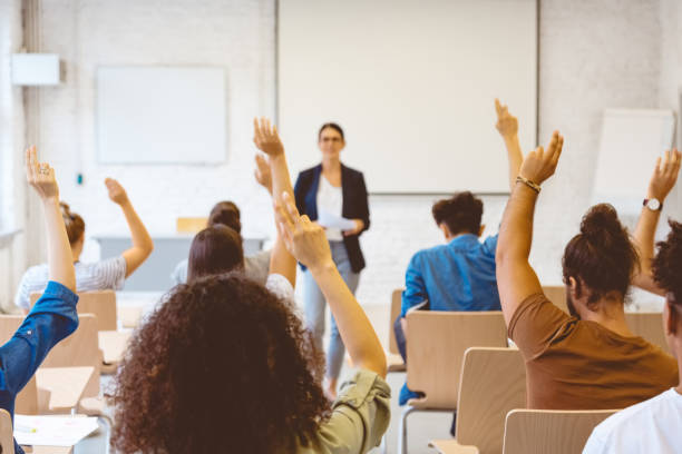 University students answering to female teacher Male and female students raising hands in classroom. Men and women are answering to mature teacher. They are together in university. lecture hall photos stock pictures, royalty-free photos & images