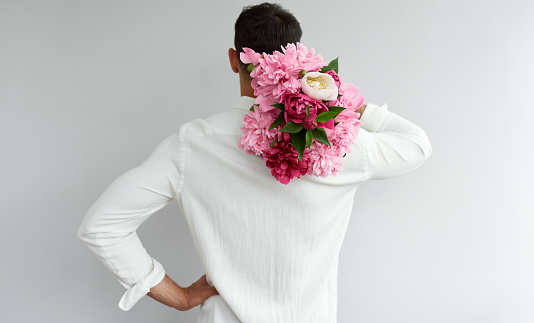 Rear view of handsome man carrying the bouquet of pink peonies as a gift for Valentine's day or wedding day. Male in white shirt holding flowers in the hands-on shoulder, isolated on the grey wall.