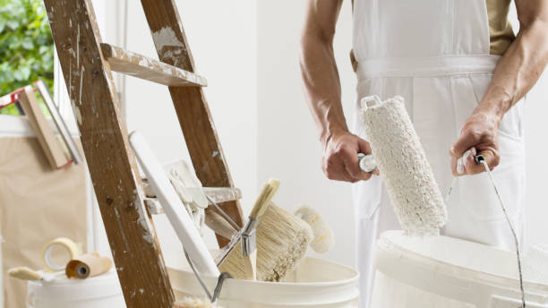 istock hands of house painter man decorator work of home to renovate, using roller paint and holding white bucket, a wooden ladder with paint brushes as background, close-up 1324199432