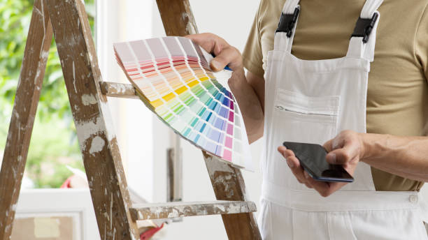 hands of house painter man decorator choose the color using the sample swatch, work of the house to renovate, use mobile phone, a wooden ladder and a green window as a background, close up - home decorator house painter color swatch paint imagens e fotografias de stock