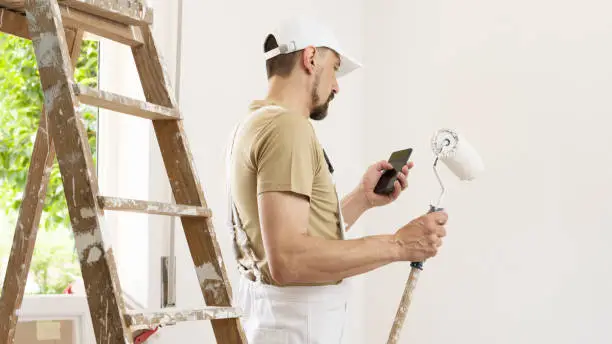 Photo of house painter man with mobile phone and paint roller, works the white home wall, a wooden ladder and green window as a background