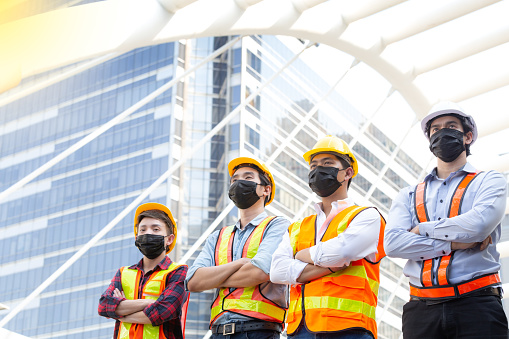 Portrait happy construction worker team with with safety helmet, safety vast reflector wearing Covid face mask standing with arms crossed on building outside. Engineering tool and construction concept