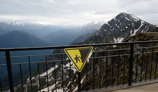 Yellow danger sign in the mountains. Warning sign of falling off cliff edge in the mountains. Danger risk of falling. Hiking Safety Warning. Danger Falling, Cliff Edge Warning Sign.