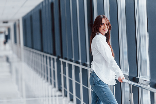 Brunette in white shirt leaning on railings indoors in modern airport or hallway at daytime.