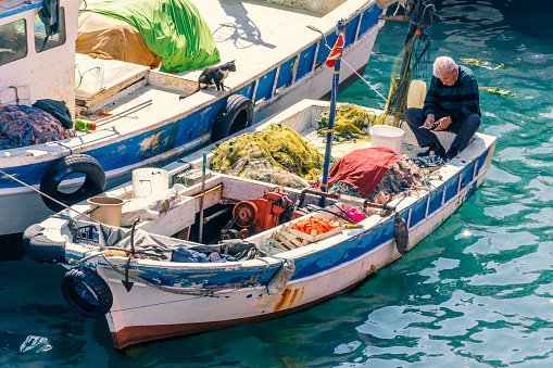 An lonely man sits in his fisher-boat at the harbor and separating the fishes from the colorful net. It is a bright day and the sun is shimmering over the turquoise water. Peaceful and typical Mediterranean background with copy space.