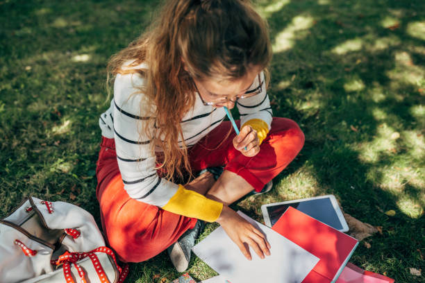 Top view of a student female in casual outfit, glasses, sitting on the green grass at the college campus, and studying with lots of books outdoors. Young woman learning and making notes in the park. stock photo